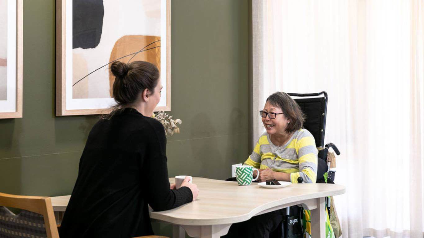 Speech Pathology Services_disability client sitting with staff member at table having a cup of tea and cake with abstract painting, khaki wall and sheers