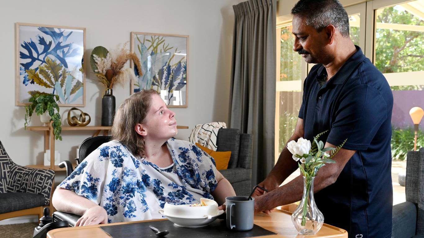 Brightwater Marangaroo Transitional Accommodation Program_disability client and disability support worker smiling together as client is sat eating meal inside